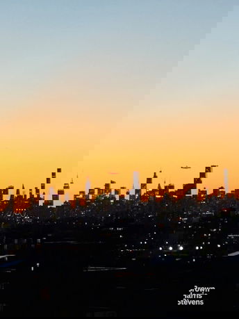 Panoramic view of a city skyline at sunset with skyscrapers and an airplane in the sky.