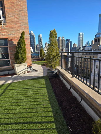 Rooftop area with artificial grass and potted plants, overlooking city buildings.