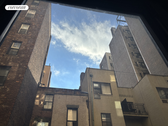 View from a window of surrounding multi-story buildings with brick exteriors under a blue sky with clouds.