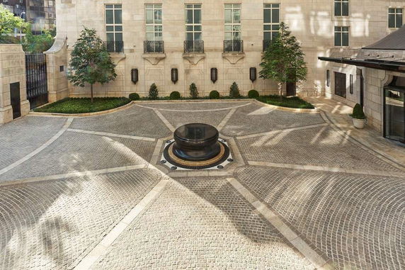 Cobbled courtyard with central round structure and surrounding greenery, adjacent to a building.