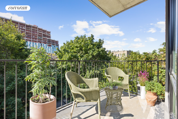 View from a balcony with wicker chairs and potted plants, overlooking trees and a partially constructed building.
