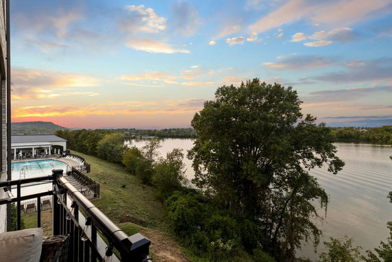 Panoramic view from a balcony overlooking a river and a swimming pool, with trees and a sunset sky.