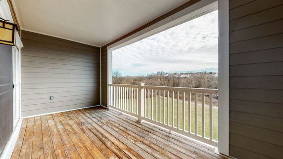 Covered outdoor balcony with wooden flooring overlooking a grassy area and distant houses.