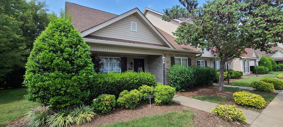 Front view of a single-story house with brick and siding exterior, surrounded by plants and trees.