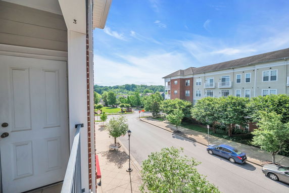 View from a building balcony overlooking a street with trees and a multi-story residential building.