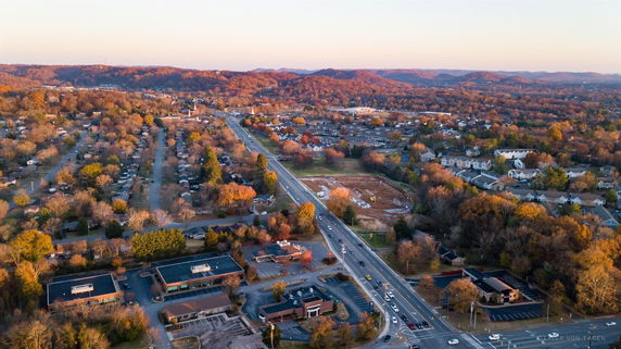 Aerial panoramic view of a suburban area with roads, houses, and trees, surrounded by hills in the background.