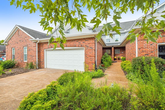Front view of a brick house with a white garage door and an entrance path surrounded by greenery.