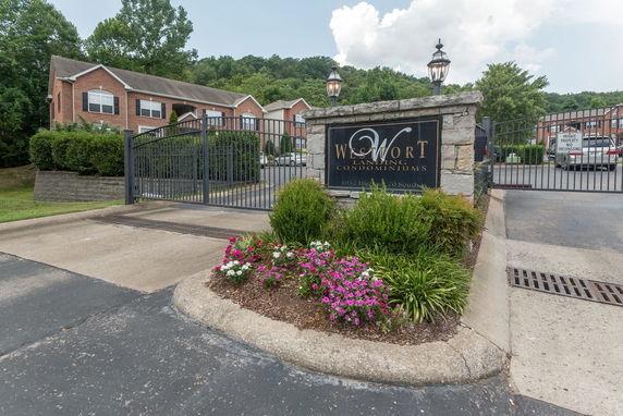 Front view of a gated condominium entrance with a sign and brick buildings in the background.